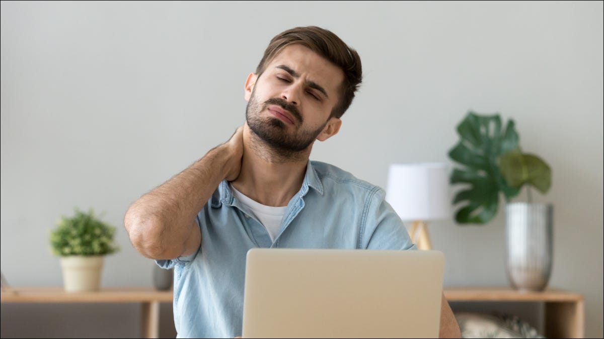 Hombre sentado frente a una computadora portátil y frotándose el cuello con dolor.